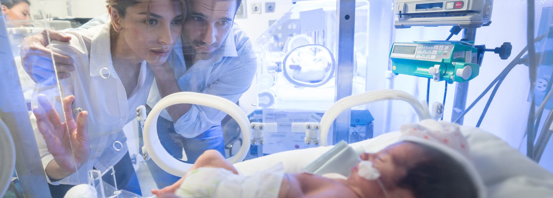 Ein Paar schaut besorgt durch das Glas eines Inkubators auf ihr neugeborenes Baby, das mit medizinischen Geräten überwacht wird. Das Baby liegt im Inkubator mit einem Sauerstoffschlauch in der Nase.