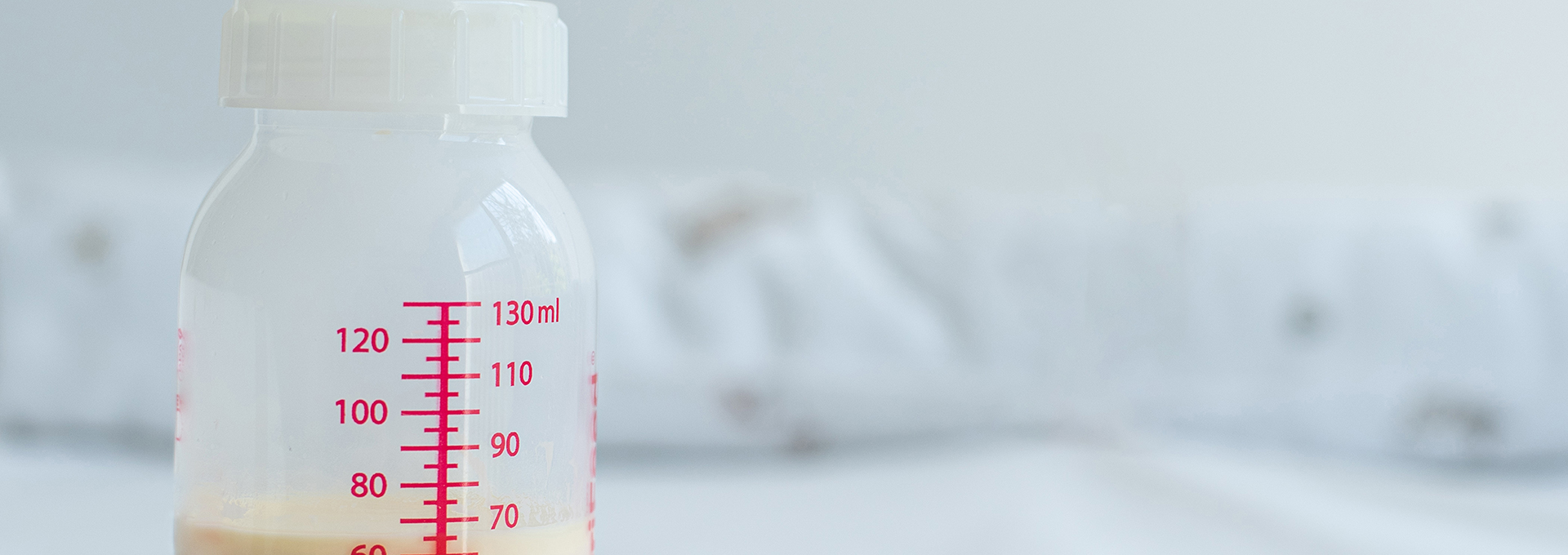 Das Bild zeigt eine Babyflasche mit einem Messskala, die bis 130 ml reicht. Die Flasche ist teilweise mit Milch gefüllt und steht auf einer weißen Oberfläche. Im Hintergrund ist ein unscharfer, heller Bereich zu sehen, der eine ruhige und saubere Umgebung andeutet. Die Flasche hat einen weißen Schraubverschluss und einen Silikonsauger, der oben sichtbar ist.