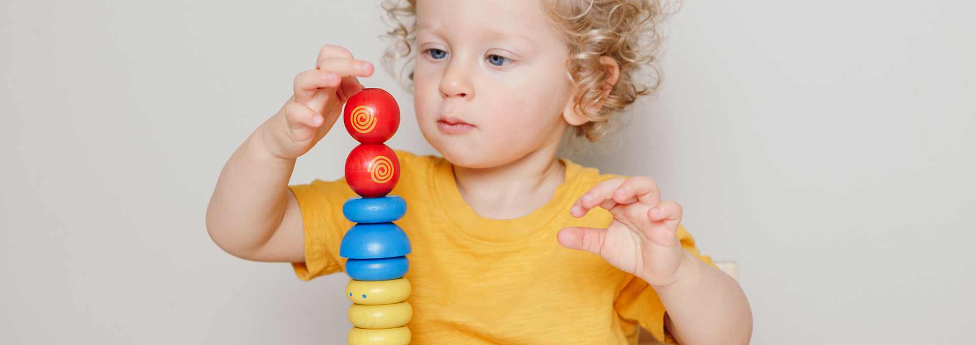 Das Bild zeigt ein kleines Kind mit lockigem blondem Haar, das ein gelbes T-Shirt trägt und konzentriert mit buntem Holzspielzeug spielt. Das Kind stapelt farbige Holzringe und Kugeln aufeinander, um einen Turm zu bauen. Auf dem Tisch vor dem Kind liegen weitere bunte Holzklötze verstreut. Der Hintergrund ist neutral und lenkt nicht von der Aktivität des Kindes ab. Das Kind wirkt vertieft und aufmerksam bei seinem Spiel.