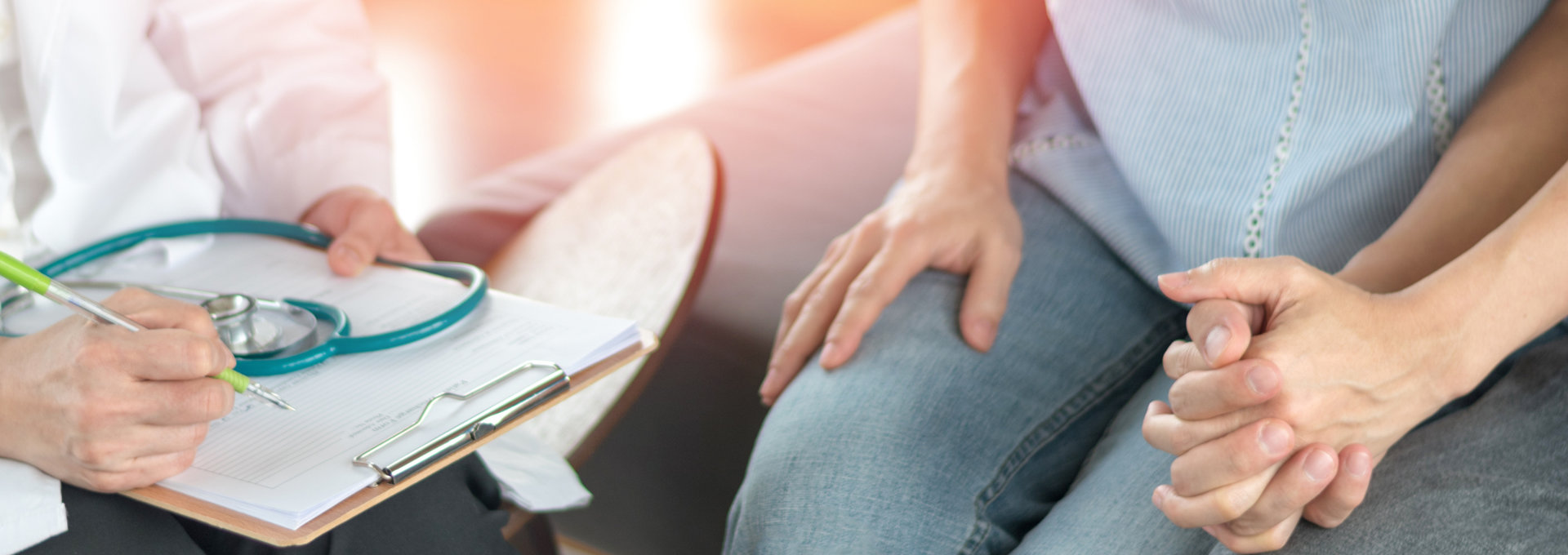 Ein Arzt sitzt mit einem Klemmbrett und einem Stethoskop auf dem Schoß und spricht mit einem Paar. Die Frau trägt ein ärmelloses Oberteil und Jeans, während der Mann eine dunkle Hose trägt. Beide sitzen eng nebeneinander und halten sich an den Händen, was auf emotionale Unterstützung und Zusammenhalt hinweist. Die Szene ist in einem hell beleuchteten Raum, möglicherweise in einer Arztpraxis oder einem Beratungszimmer, und vermittelt eine Atmosphäre der Kommunikation und des Vertrauens zwischen dem Arzt und den Patienten.