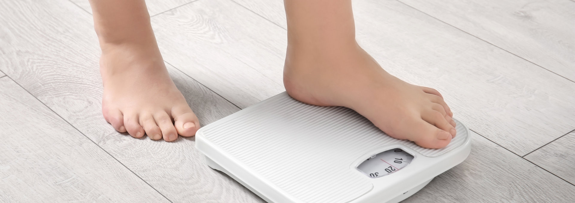 Eine Person steigt mit einem Fuß auf eine weiße digitale Badezimmerwaage, während der andere Fuß noch auf dem Boden steht. Die Szene spielt sich auf einem hellen Holzfußboden ab, und die unteren Beine und Füße der Person sind sichtbar, was darauf hinweist, dass sie ihr Gewicht überprüft.