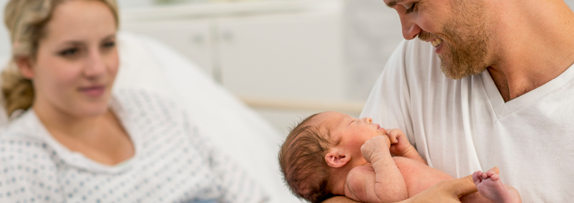 Ein Vater hält sein Baby in den Armen. Im Hintergrund liegt die Mutter auf einem Krankenbett.