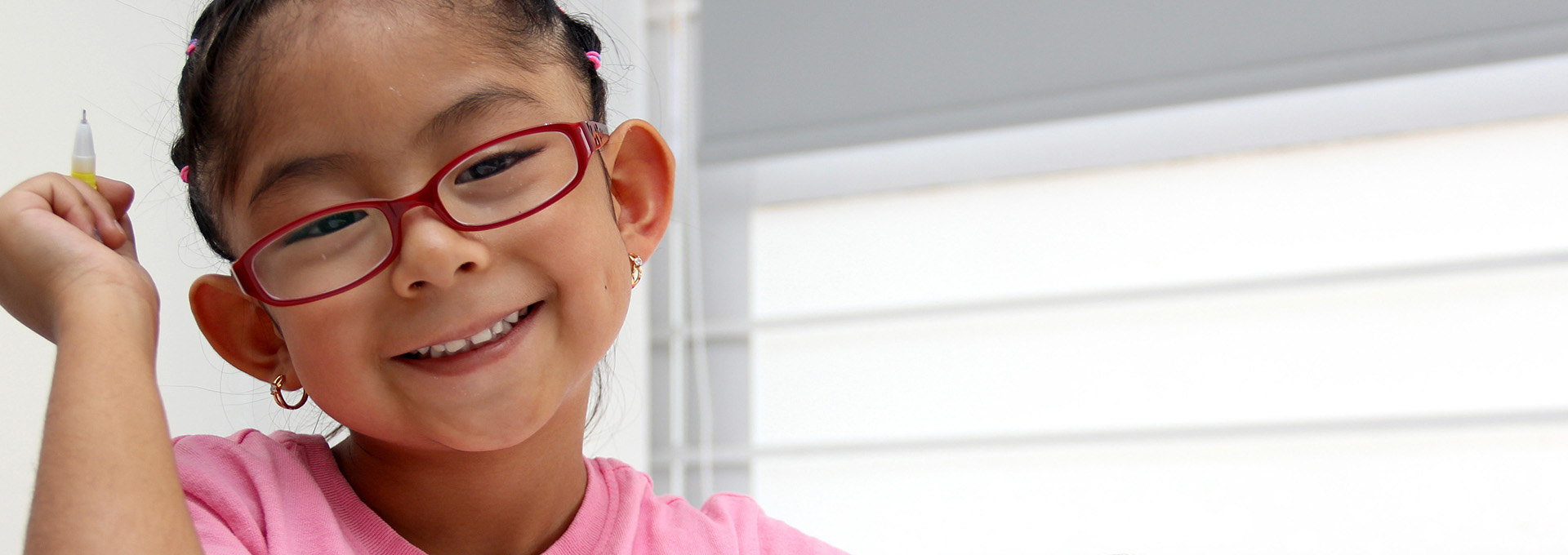 Ein Mädchen mit Brille und pinken Shirt hält einen Stift in der Hand und lächelt.