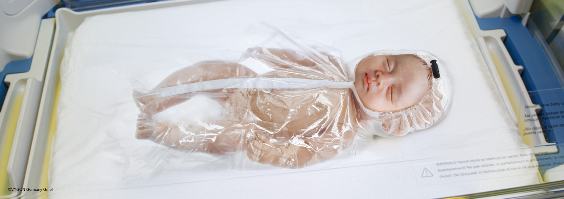 Ein Baby, das in eine Plastikfolie gewickelt in einem Brutkasten liegt.