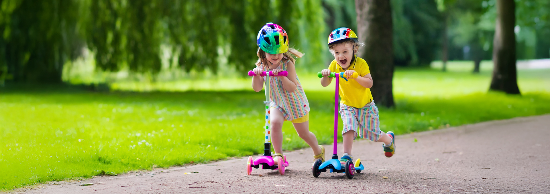 Zwei Kinder mit bunten Helmen fahren lachend auf Tretrollern in einem Park.
