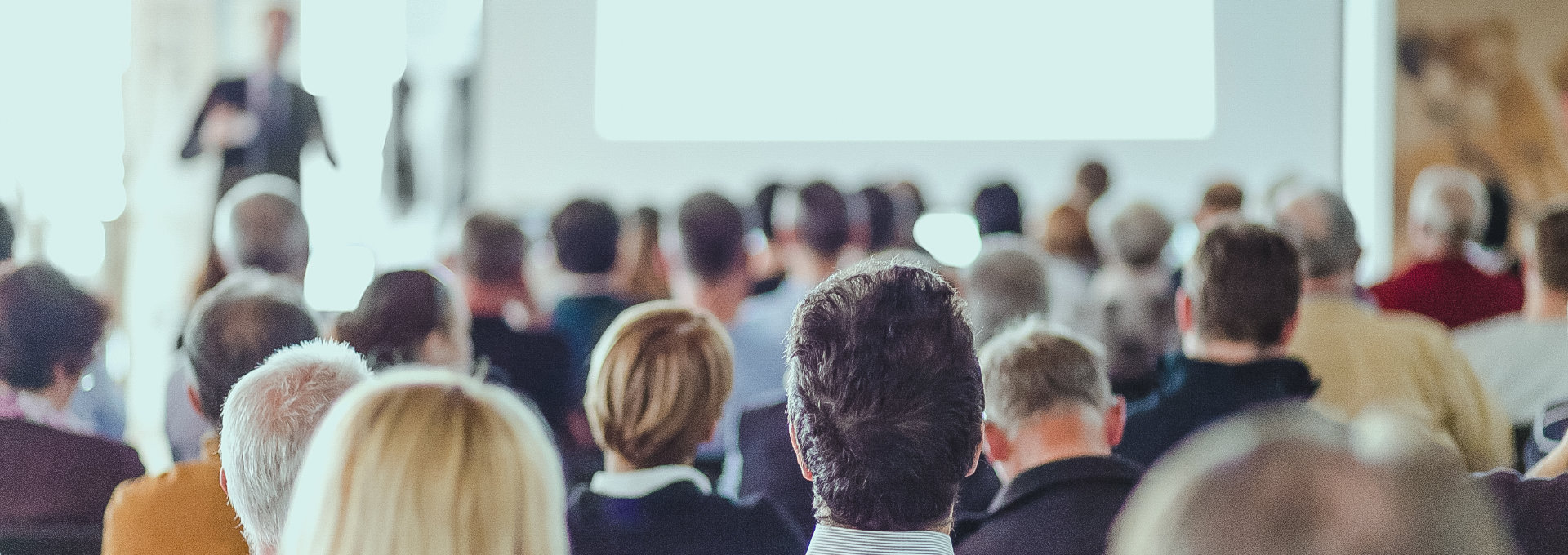 Ein voller Konferenzsaal und das Publikum schaut auf einen Vortragenden, der unscharf zu erkennen ist. Man erkennt nur die Rücken der Personen.