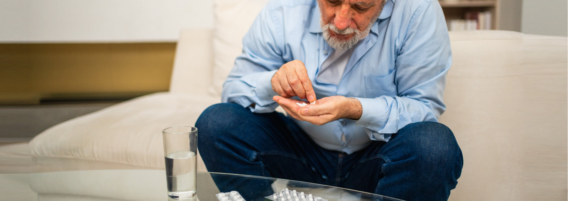 ein älterer Mann sitzt mit grauem Bart sitzt auf einem beigefarbenen Sofa und zählt in seinen Händen weiße Tabletten ab, vor ihm auf dem Tisch steht ein Wasserglas und liegen zwei Tabletten-Blister