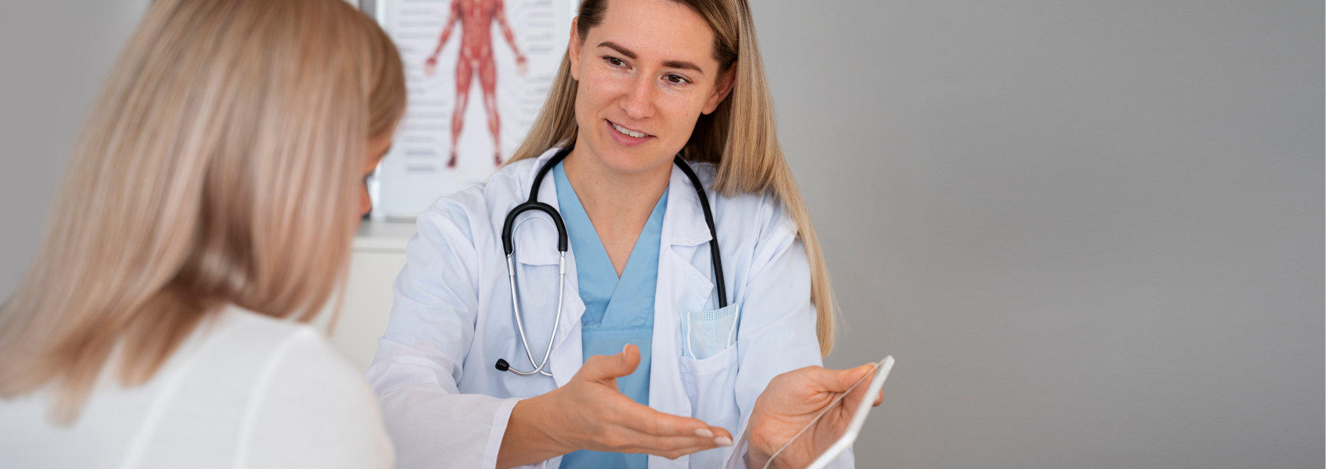Junge blonde Ärztin sitzt am Schreibtisch und erklärt einer Patientin etwas anhand eines Tablets