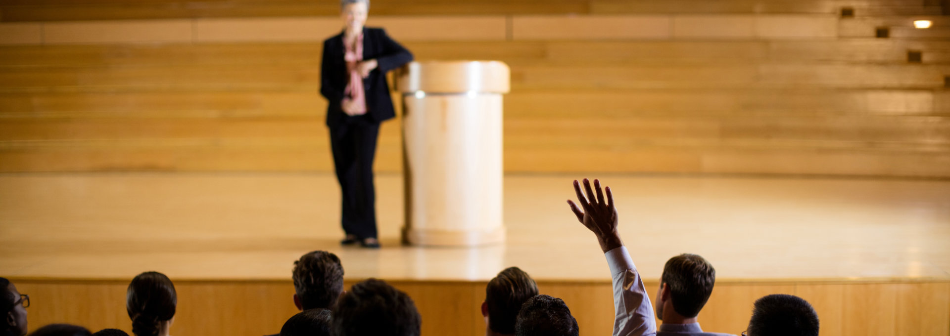 eine Frau steht auf einem Podium an ein Rednerput gelehnt, im Vordergrund sind Köpfe des Publikums zu sehen und eine Hand, die sich meldet