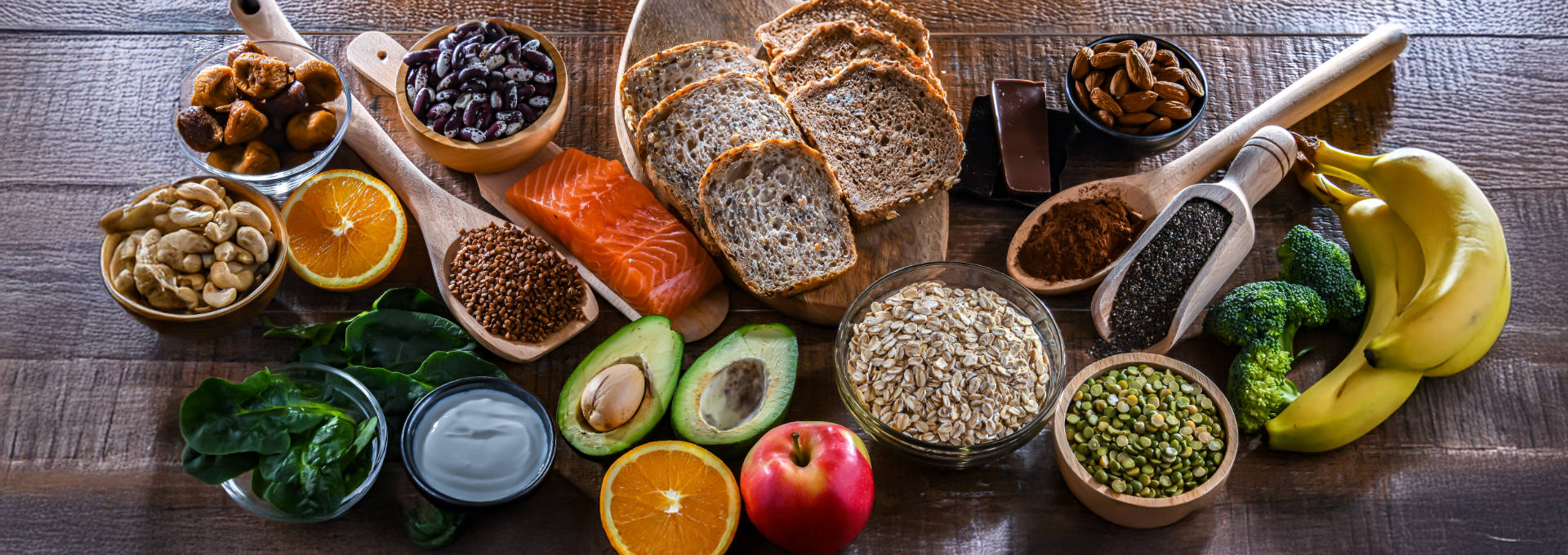 Eine Auswahl an gesunden Lebensmitteln auf einem Holztisch, darunter Vollkornbrot, Lachs, Avocado, Obst, Gemüse, Nüsse, Hülsenfrüchte und Haferbrei.