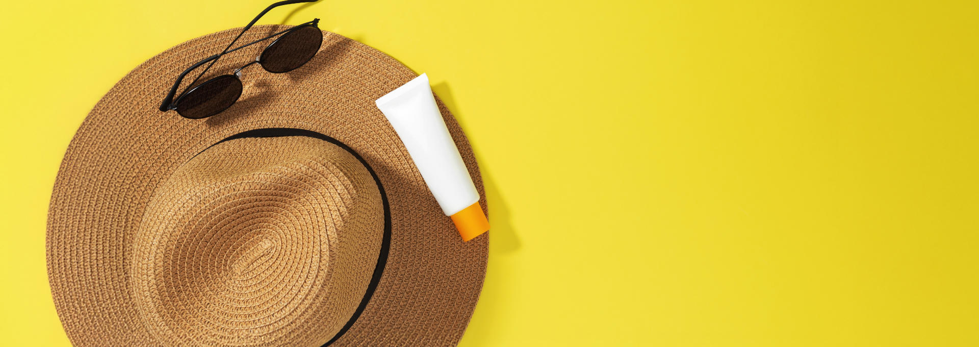 Strohhut, Sonnenbrille und Sonnencreme auf gelbem Hintergrund.