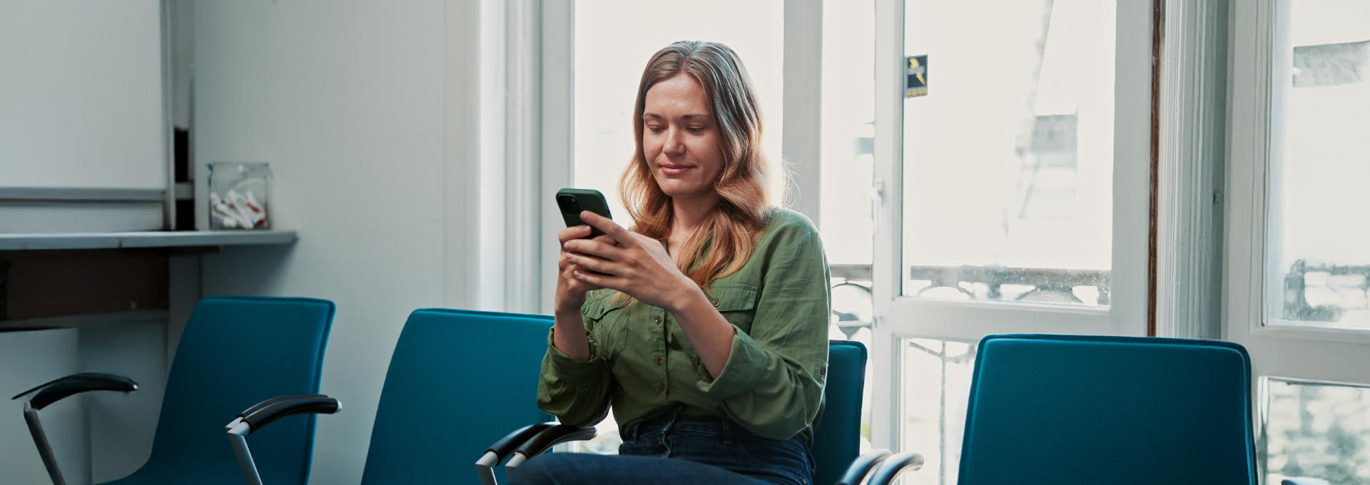 Eine junge Frau sitzt in einem Wartezimmer und schaut auf ihr Smartphone