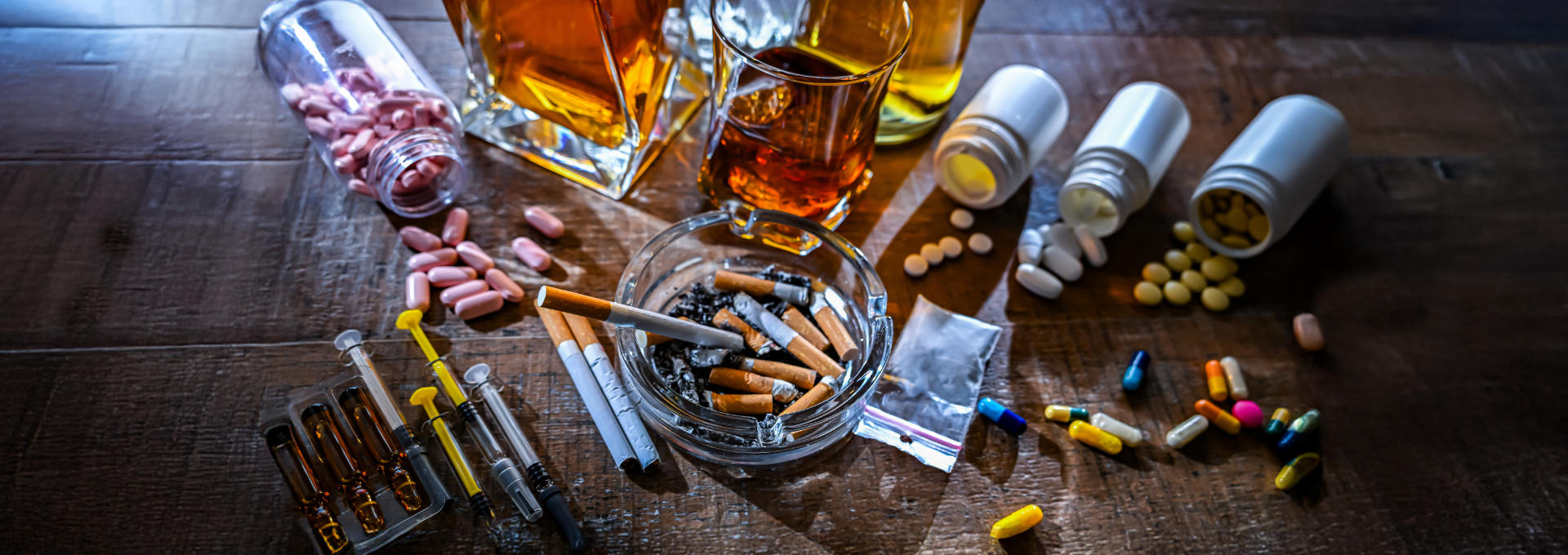 Auf einem Tisch sind ein Aschenbecher mit Zigaretten, Alkohol in Glasbehältern, Pillen und Spritzen ausgebreitet