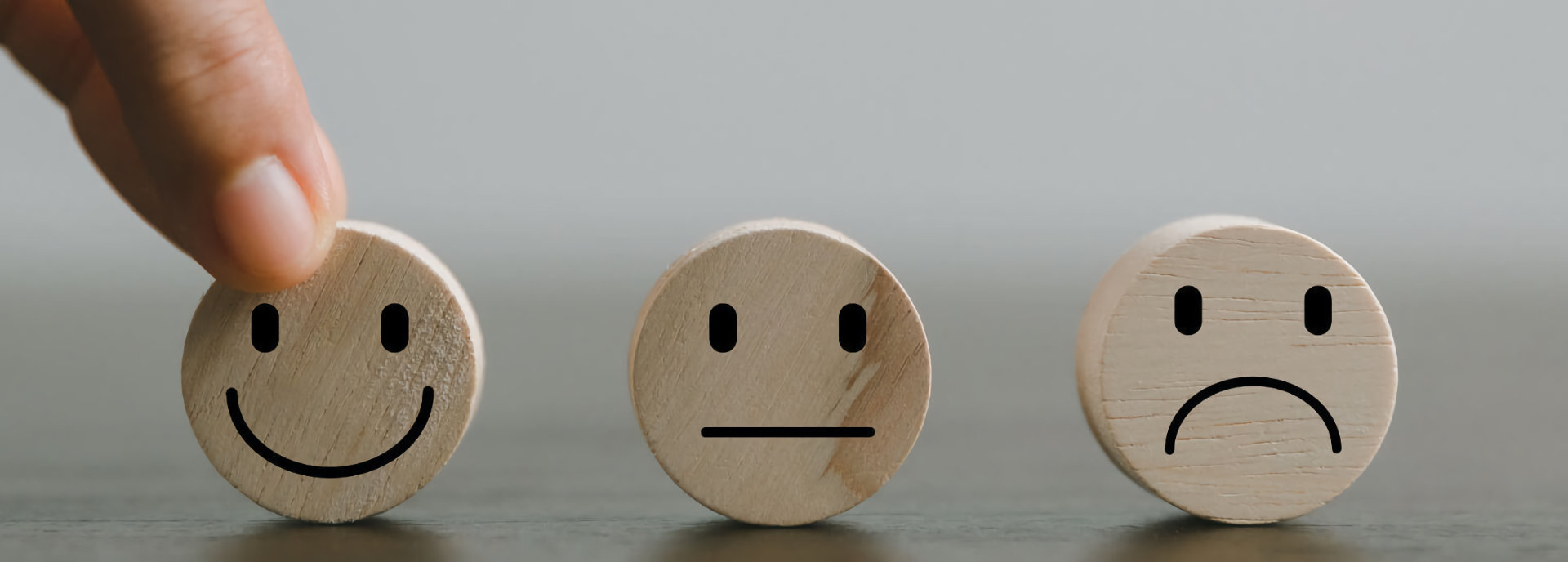Ein Bild zeigt drei runde Holzscheiben mit aufgemalten Gesichtern, die verschiedene Emotionen darstellen. Von links nach rechts: ein lächelndes Gesicht, ein neutrales Gesicht und ein trauriges Gesicht. Eine Hand wählt die lächelnde Holzscheibe aus, was einen positiven Ausdruck hervorhebt. Der Hintergrund ist schlicht und grau.