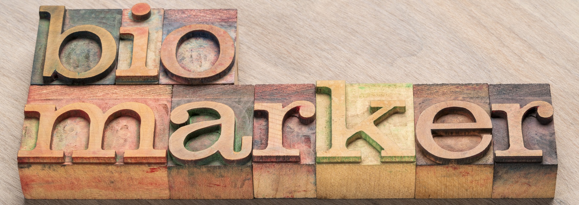 Holzbuchstaben formen das Wort „biomarker“ auf einer hellen Holzoberfläche. Die Buchstaben sind in verschiedenen Farben und leicht verwittertem Stil gestaltet.