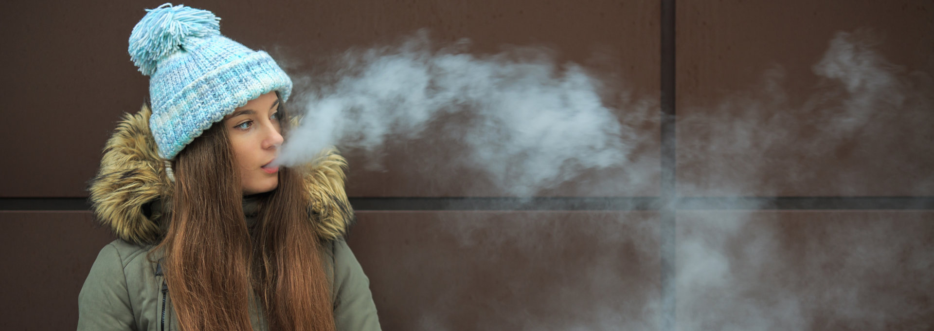 Junge Frau mit langen, braunen Haaren, die eine blaue Mütze und eine grüne Winterjacke mit Fellkragen trägt, bläst beim Vapen eine große Wolke Dampf aus. Sie steht draußen vor einer dunkelbraunen Wand