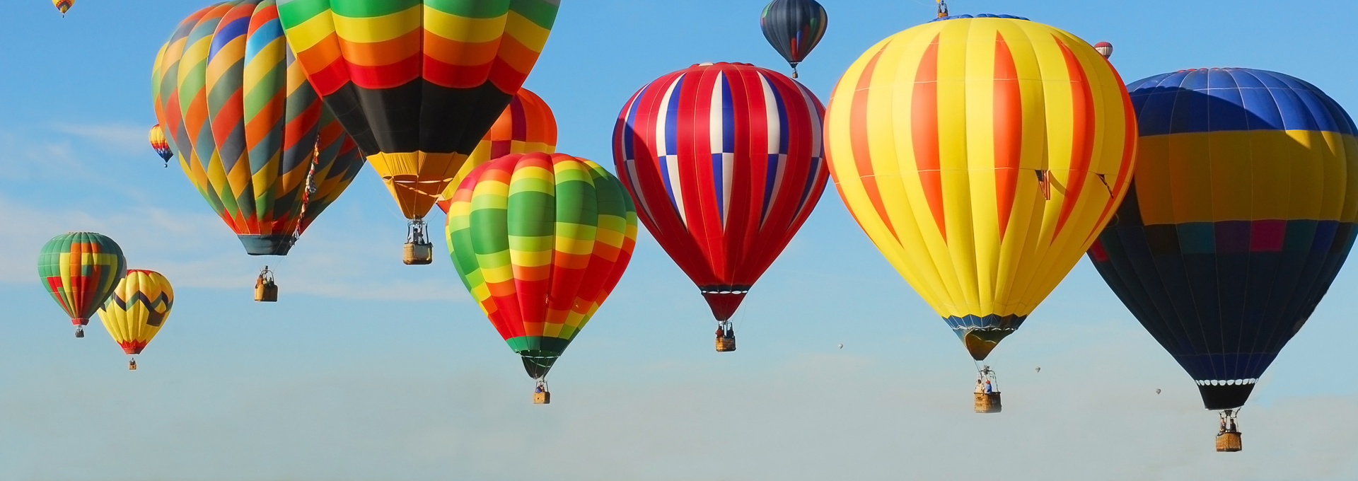 Mehrere bunte Heißluftballons schweben bei klarem, blauem Himmel in der Luft. Die Ballons haben unterschiedliche Farben und Muster, darunter Streifen und Rauten.