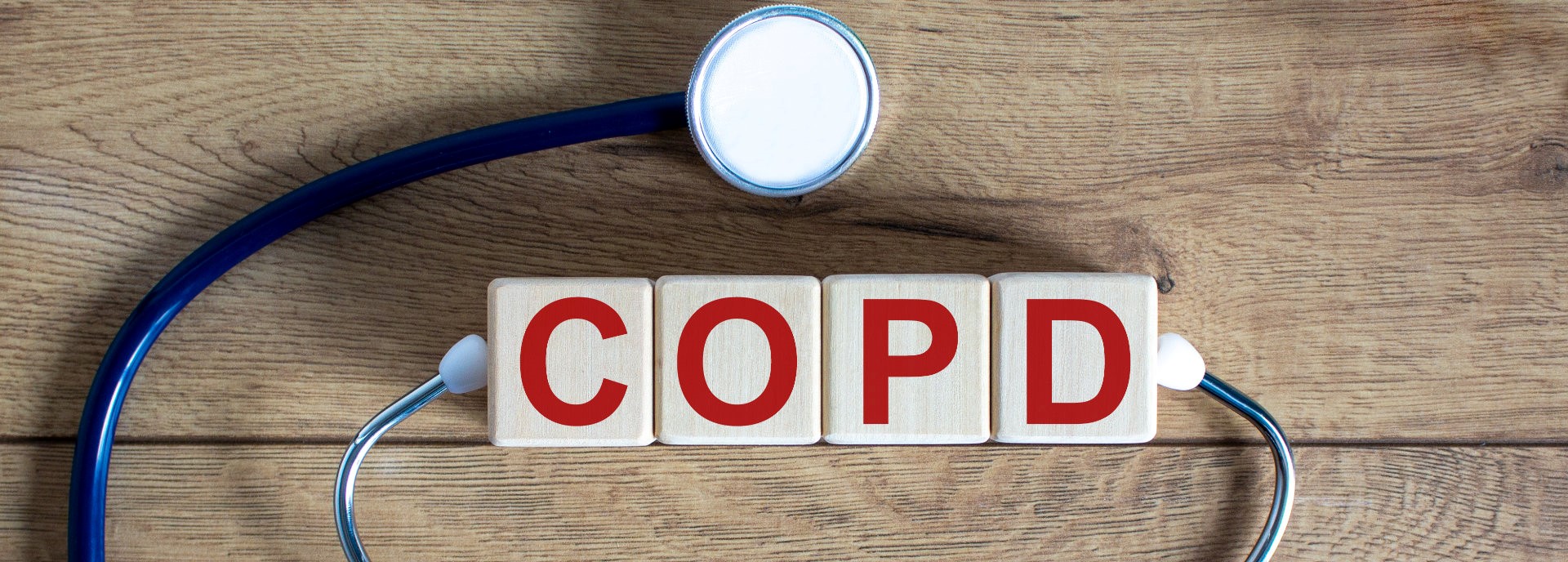 Ein Bild zeigt einen hölzernen Schreibtisch, auf dem Holzklötze mit den Buchstaben "COPD" in roter Farbe angeordnet sind. Um die Klötze ist ein Stethoskop mit blauem Schlauch drapiert, das den Text umschließt. Der Hintergrund ist die Holzoberfläche des Tisches